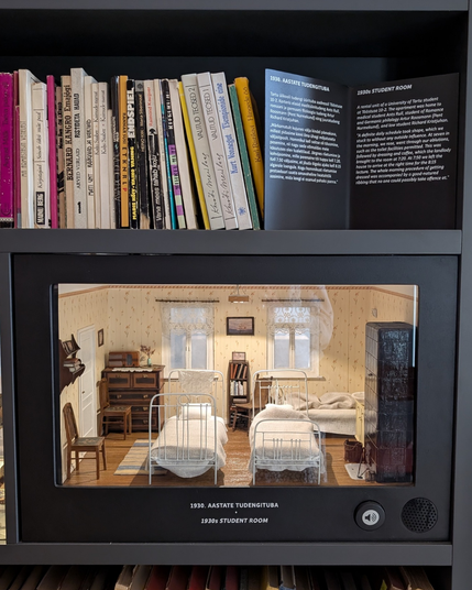 Grāmatplaukts ar 1930. gadu studentu istabas miniatūru attēlojumu.