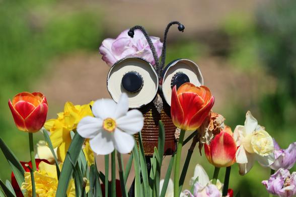 Ein braunes Insekt inmitten von Tulpen und Narzissen. Der Körper eine verrostete Blechdose, die Augen zwei Deckel von Schraubgläsern mit Kronkorken als Pupillen, zwei Fühler aus Bürstendraht mit je einer Perle am Ende.