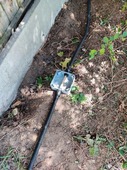 Grey junction box on top of dirt. Black conduit leads in to and out of it, with some grey and orange cables running through the middle.