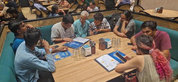 Agonizing over what to order.

7 men and a girl sitting on green sofas around a wooden table, poring over menus in a variety of attitudes.