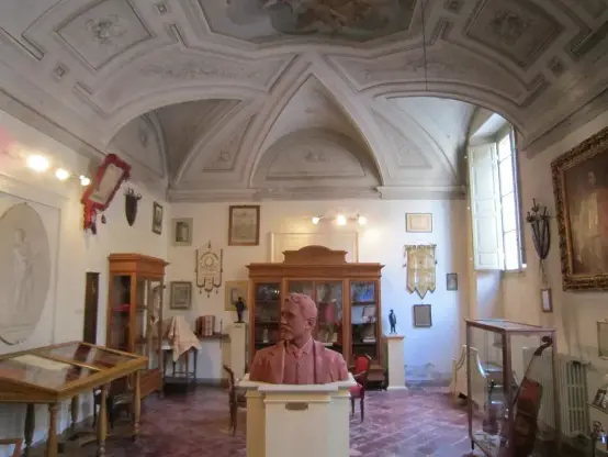 The image depicts an interior of a museum or historical room. It features a bust of a man, likely displayed on a pedestal in the center. Surrounding the bust are various exhibits, including cabinets and other artifacts. The ceiling is adorned with decorative elements, and there are windows allowing natural light to enter the space. The overall atmosphere suggests a place dedicated to history or art.

Image Credits: Wikimedia / Perkele / Public domain