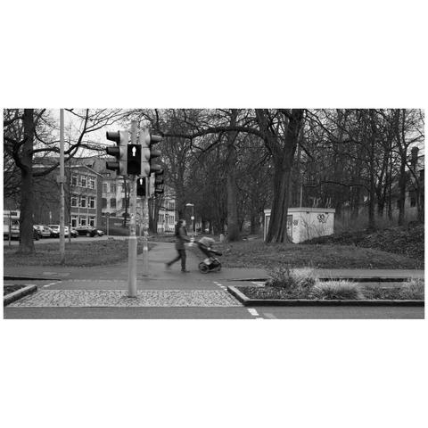 Picture of a pedestrian crossing with red light. On the opposite side a person walks by with a pram, blurred slightly due to the slow shutter speed.