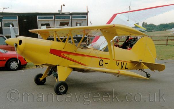 Side view of a yellow, single-engined bi-plane parked facing to the left.
There are a pair of tandem open cockpits, with glazed windshields in front of each.
There is a red stripe running along the body from just behind the engine, also heading to the lower rear fuselage.
Black tarmac fills the foreground, with the wing and fuselage of a microlight behind this plane on the right, and a pair of red or orange cars on the left.
A black, pre-fabricated building fills the background on the left of the frame, with hazy grey sky filling the rest.
