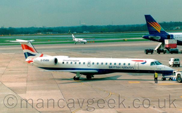 Side view of a white and blue, twin engined jet airliner with the engines mounted on the sides of the rear fuselage, facing to the right but being pushed back to the left by a low-slung white and blue tug attached to the nose-wheel by a long blue pole.
The plane is mostly white, with a blue belly, and blue "British Airways" titles on the lower forward fuselage, just aft of the forward door and below the passenger cabin windows.
There is an image of a 2-sided, twisted red and blue ribbon on the upper forward fuselage, above the forward door.
There are black "United Kingdom" titles on the upper rear fuselage, and the registration "G-EMBN" on the side of the engine pod.
There are wavy red and blue lines on the tail.
A man wearing a hi-viz jacket and a pair of rear ear defenders is walking alongside the plane as it is pushed back.
Grey concrete apron fills the foreground, with more in the background, leading up to grass and taxiways in the distance, with trees and the dish of a radio-telescope right at the back, slowly being swallowed up in hazy sky.
The dark blue tail of aa larger jet airliner is poking into the frame from the right, with a smaller, white, twin propellor-engined airliner in the middle of the frame.