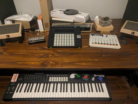 Escritorio con bandeja retráctil con un novation launchkey 61 mkii, arriba un push 2, un volca bass, un po-20, arturia drumbrute impact 1984 y dos soportes de parlantes rodeados de cajas.