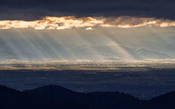 Blick vom Schwarzwald über die Rheinebene zu den Vogesen, mit Teleobjektiv aufgenommen. Den Vordergrund bilden dunkle Schwarzwaldhügel, die im Schatten liegen. Dahinter liegt die Rheinebene, die in zwei kontrastierende Hälften geteilt ist: vorne im Schatten, hinten sonnendurchflutet. Darüber sind die Hügel der Vogesen im Dunst zu erkennen. Am oberen Bildrand hängt eine dunkle Wolkendecke, deren unterer Rand von hinten durch die Sonne angestrahlt ist. Das Sonnenlicht, das die Landschaft darunter beleuchtet, bildet ausgeprägte Strahlen im Dunst.