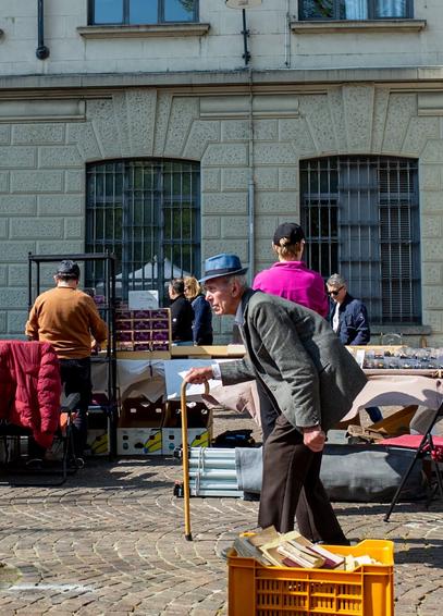 The photo shows an old gentleman walking between flea market stalls with his back bent. He is dressed for Sunday, wearing a suit and hat and leaning on an old-fashioned cane.
