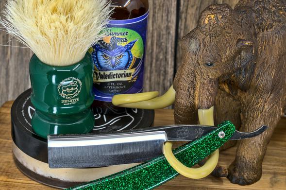 (Le Chat Mistral description)

This image features a collection of grooming items arranged on a wooden surface. There's a green shaving brush with a white bristle head, placed in a matching green holder. Next to it is a bottle of aftershave labelled "Summer Break Valedictorian," with a blue and green design featuring an owl. In front, there's a straight razor with a green glittery handle and a silver blade. To the right, there's a small figurine of a mammoth with curved tusks. The background includes a wooden texture, adding a rustic feel to the scene.