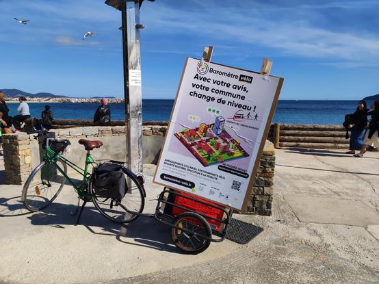 Photo d'un petit vélo vert de ville, avec saccorche et remorque, garé à la Plage du Mourrillon. La remorque porte une énorme affiche (format A0) avec le visuel du baromètre vélo, ainsi que lien et logo de TVD.