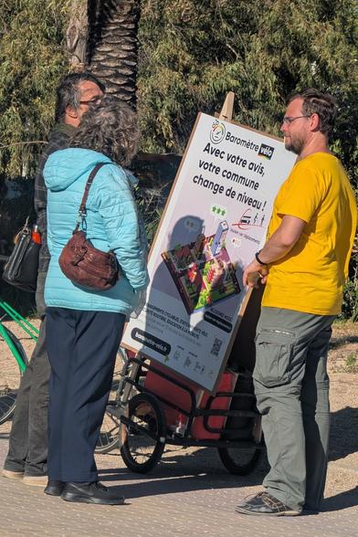 Photo d'un homme blanc parlant avec un couple de vieux homme/femme blancs. Les trois sont debout à côté de la remorque vélo de l'autre photo.