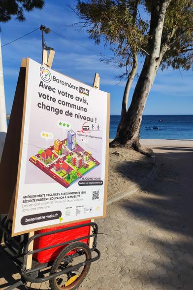 Photo d'une remorque vélo garé à la plage du Mourillon. Elle porte une énorme affiche (format A0) avec le visuel du baromètre vélo, plus le logo et le lien de TVD.