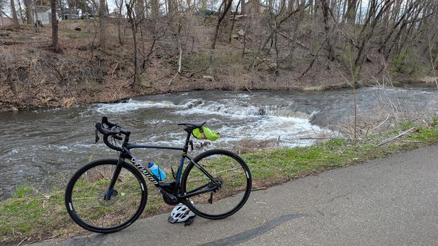 Blue and black Roubaix bicycle parked on the Cascade Creek Trail with a cascade of the creek behind, the water is high and flowing over all of the falls, greening grass on this side and mostly bare trees and dead leaves on the ground on the opposite side of the creek.