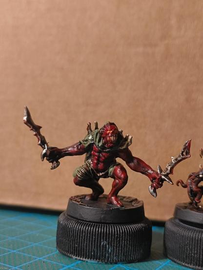 A red demon in goblin squad pose, dressed in green rags and armed with two swords.