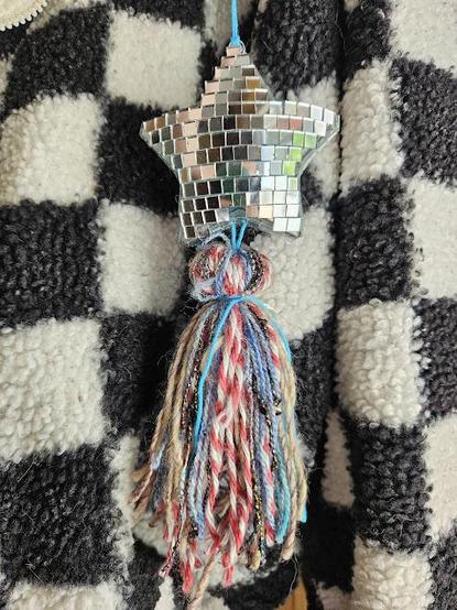 A star-shaped ornament covered in mirror tiles to look like a disco ball, with a multicolored yarn tassel hanging from the bottom, in front of a black and white checkered background.