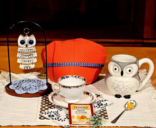 Tea tableau with owls. There are a figural owl shaped mug, a jointed metal owl, vintage china, a dark orange cozy, a spoon with oranges painted on it, and a packet of Orange Peach herbal tea from Twinings