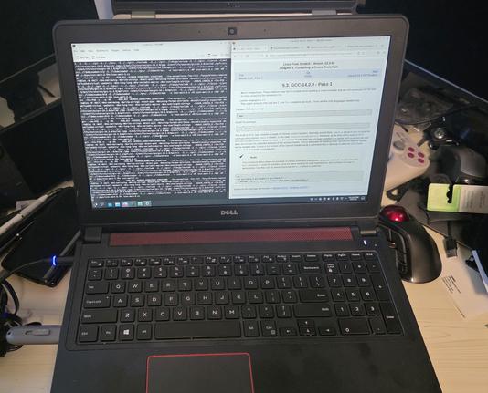 Dell laptop sitting on a cluttered desk. On the screen are two windows opened, one side is a terminal compiling GCC from source, the other is a browser opened to the Linux From Scratch documentation.