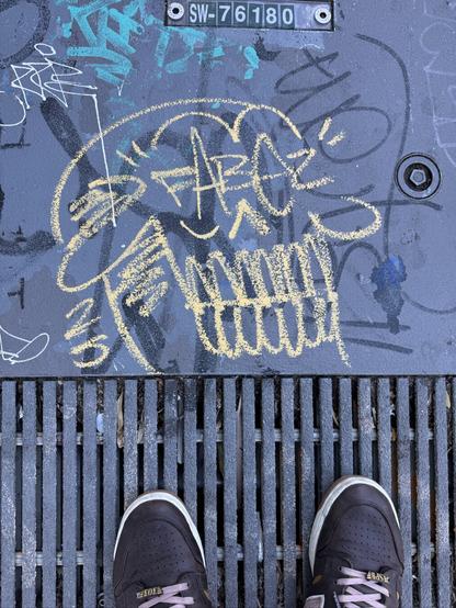 A skull drawn in graffiti style on the sidewalk. The skull has a big toothy grin and tiny wings growing out of the top. It says “Fabez”, the artist’s name, on the skull where the eyes would normally be.