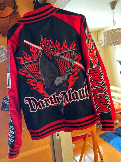 An embellished black and red bomber jacket featuring a large graphic of a character with a red and black mask, wielding a lightsaber amidst flames. The back displays the text "Darth Maul." The jacket has striped cuffs and various designs.