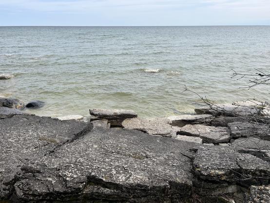 A photo of dark rocks that are up against a grey green/blue lake with a blue sky above it.