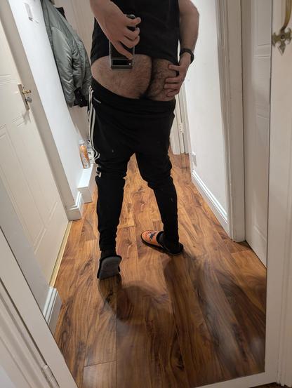A man taking a selfie of his back, with his tracksuit pants down so his hairy ass is on view. He's also wearing a dark t-shirt and Bert and Ernie slippers