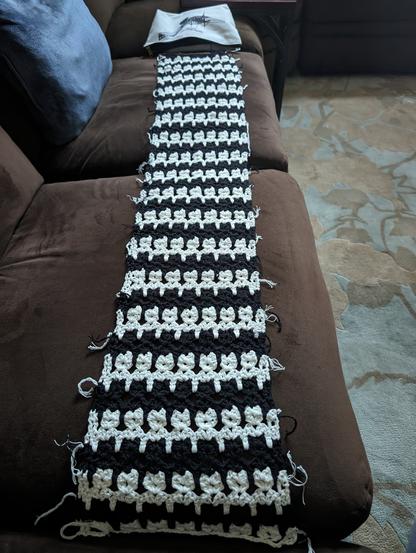 a black-and-white scarf in repeating rows of tessellated cat stitch, with loose ends every row waiting to be worked in, on a brown sectional sofa