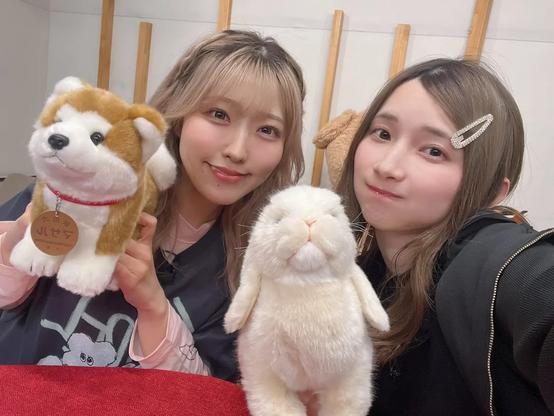 Two young women smile at the camera while holding up plush toys—one of a Shiba Inu dog and the other of a white bunny—creating a cute and cozy atmosphere in a warmly lit indoor setting.