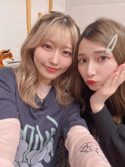 Two young women pose closely for a cheerful selfie, one smiling sweetly and the other making a playful pout, with a stuffed fox toy visible in the background on a shelf.