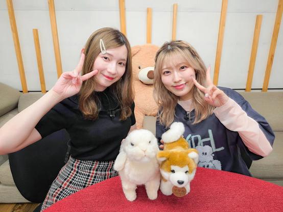 Two young women smile and pose with peace signs while sitting at a table with plush toys of a white rabbit and a Shiba Inu dog, with a large teddy bear in the background on a cozy, modern couch.