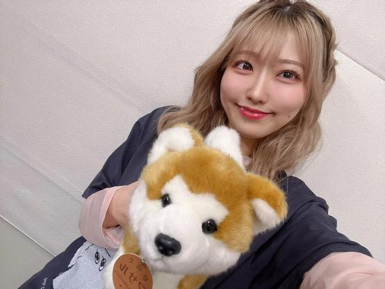 A young woman smiles brightly while holding a plush Shiba Inu toy close to the camera, with a soft neutral background enhancing the cozy, cheerful vibe of the photo.