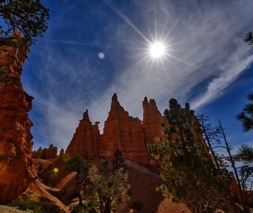 A picture in the Queen's Garden, Bryce National Park, where one of the hoodoos bears a striking similarity to Queen Victoria - apparently.  Other hoodos in this picture too, one on the left foreground, more in the middle distance with QV the left most.  Blue sky with wispy coulds, and the sun in frame showing a pattern of lines going out from it - shot at small aperture to get the lines.
