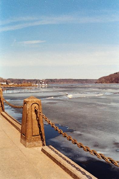 View of the partially frozen St. Croix River in Stillwater, Minnesota. A concrete and chain railing runs along the riverside path. The ice is beginning to break up under a pale blue sky, suggesting the end of winter. Shot on film.