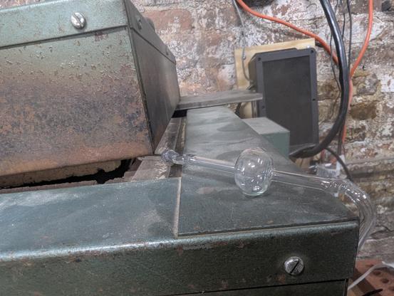 A glass tube with a bubble in it emerging from a crack in a kiln oven door. The door is propped open with a metal plate in the back. There is a small ball of mercury sitting in the bottom of the bubble.