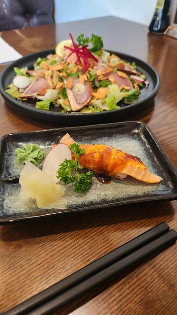 A small plate with a salmon aburia nigiri and garnishes. Behind it is a large plate with salad, daikon and tuna tataki topped with a beet garnish.