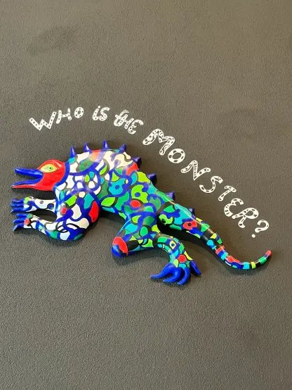 Une sculpture de lézard colorée et stylisée est exposée sur un fond gris texturé. Au-dessus du lézard, le texte "Qui est le MONSTRE ?" Est écrit artistiquement.