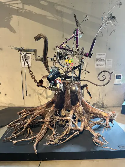 Une sculpture en mosaïque colorée ressemblant à une grande forme abstraite est exposée à l'intérieur. Il présente un design bleu et vert avec des accents miroir et un personnage ludique représenté d'un côté. L'espace environnant comprend des éléments architecturaux modernes et des œuvres d'art supplémentaires en arrière-plan. Les signes indiquent