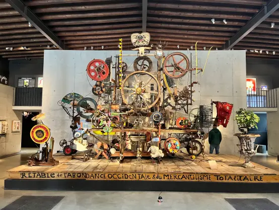 Une installation artistique élaborée à plusieurs niveaux présentant divers objets trouvés, des roues et des décorations colorées, intitulée "Retable de l'Abondance Occidentale & du Mercantilisme Totalitaire".