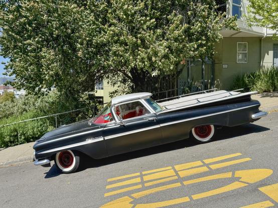 Photograph from a side profile view of a vintage #Chevrolet #ElCamino automobile. The vehicle’s space-age styling is accentuated by its jutting fins, decorative chrome trim and cargo rails, red interior dashboard, and red-hubbed whitewall tires. The car is painted a matte black with few blemishes. The vehicle is parked on a steep incline at the curb of an urban street with lush green trees and a green-painted house visible in the background.