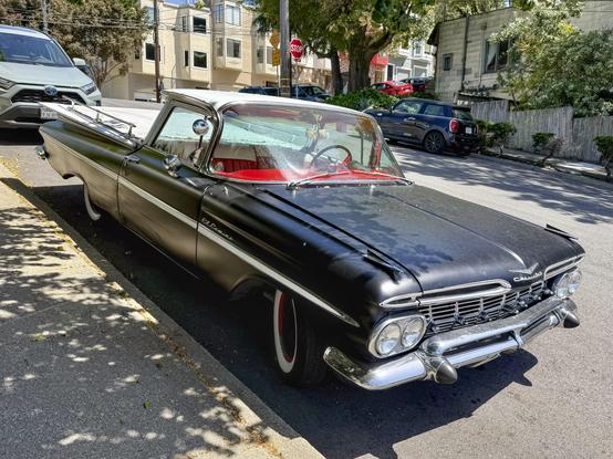 Photograph from a front three-quarters view of of a vintage #Chevrolet #ElCamino automobile. The camera’s perspective accentuates its bulbous glass windshield, red interior dashboard, white-painted running stripe, and extravagantly chromed grille and bumper. The car is painted a matte black with few blemishes. The vehicle is parked on an incline at the curb of an urban street with other cars, trees, and mid-height houses visible in the background.