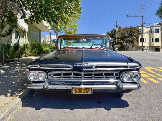 Photograph from a head-on front view of of a vintage #Chevrolet #ElCamino automobile. The camera’s perspective accentuates its wide hood, stylized script Chevrolet badge, tapered air intakes, four round headlights, and extravagantly chromed grille and bumper. The car is painted a matte black with few blemishes. The vehicle is parked on an incline at the curb of an urban street with trees and mid-height houses visible in the background.