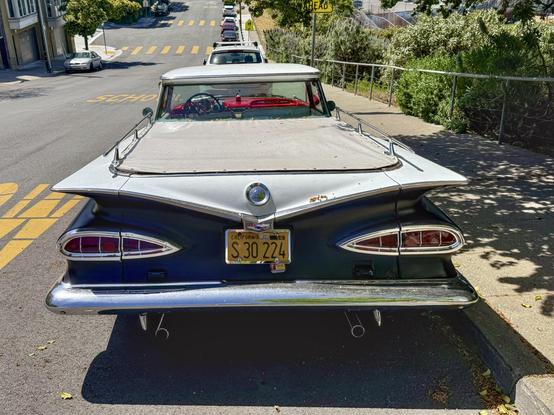 Photograph from a direct rear view of of a vintage #Chevrolet #ElCamino automobile.The camera’s perspective accentuates the space-age styling of the vehicle’s tarpaulin-covered cargo bed, chrome cargo rails, splayed fins, cat-eye taillights, and dual exhaust pipes. The car is painted a matte black with few blemishes. The vehicle is parked on an incline at the curb of an urban street with green shrubbery and other parked vehicles visible in the background.