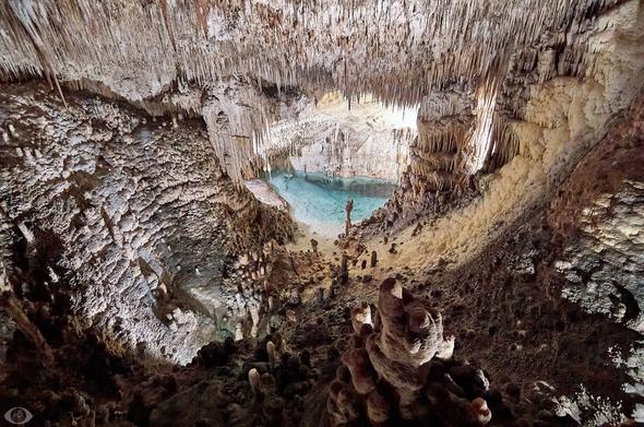 Bird’s eye view of a subterranean lake in the caves of cuevas darch (Mallorca.) Lots of long and small stalactites hanging from the ceiling of the cave with more widely stalagmites in the foreground.