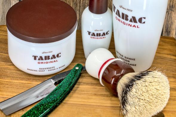 (Le Chat Mistral description)

This image shows a collection of men's grooming products arranged on a wooden surface. There is a white jar of shaving soap with a brown lid, labelled "Tabac Original" since 1959. Next to it is a white bottle of aftershave lotion with a similar label. A shaving brush with a brown handle and white bristles is placed nearby. Additionally, there is a straight razor with a green glittery handle and a silver blade. The items are neatly displayed, suggesting a classic shaving setup.
