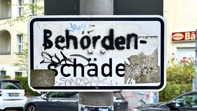An urban street sign partially obscured by graffiti. The visible text reads “Behördenschade,” while the sign shows signs of wear, including scratches and a faded area. In the background, there are parked cars and a building with greenery.