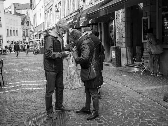 The image depicts a black and white scene of a cobblestone street in Bruges, Belgium. In the foreground, two individuals are engaged in a conversation. The person on the left is wearing a dark jacket, jeans, and glasses, holding a small object, possibly a receipt or a card. The person on the right is dressed in a long coat, holding a patterned shopping bag, and appears to be showing something to the other person.