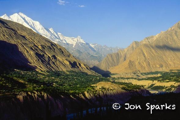 A valley with green irrigated terraces set against steep barren slopes is backed by a dominant snowy mountain peak.