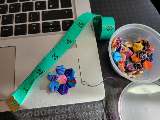 Photograph of my pot of tiny paper pieced fabric hexagons alongside a flower made by sewing seven of them together, with a green tape measure and slim needle threaded with purple thread. The flower is predominantly blue with pink red, yellow and green and has been whip stitched together with purple thread.  

The pot contains a load of multicoloured hexagons which have been thread basted. The flower, needle and tape measure are sitting on my laptop, because the magnet of the laptop is keeping the tape measure in place.