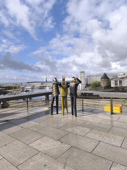 Das Bild zeigt drei große, stilisierte Figuren aus Metall auf einem gepflasterten Platz mit Blick auf den Hafen von Brest. Die Figuren sind in verschiedenen Farben gehalten: eine ist gelb mit schwarzen Streifen, eine ist schwarz und eine ist goldfarben. Sie stehen nebeneinander, mit dem Rücken zum Hafen, wobei die goldene Figur die Hand zum Gruß hebt. Im Hintergrund ist der Hafen mit Booten und einigen Gebäuden zu sehen, darunter der Turm Tanguy. Der Himmel ist teilweise bewölkt.