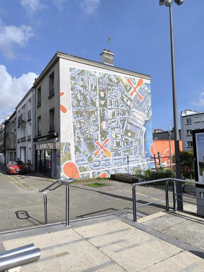 Das Bild zeigt ein Gebäude mit einer großen Wandmalerei, die eine Stadtplanansicht darstellt. Die Wandmalerei ist in verschiedenen Farben gehalten, hauptsächlich in Blau, Grau und Orange, und zeigt Straßen, Plätze und Gebäude in einer stilisierten Form. Auf der Wand sind mehrere große, orangefarbene "X"-Symbole verteilt. Der Himmel ist blau mit einigen weißen Wolken.