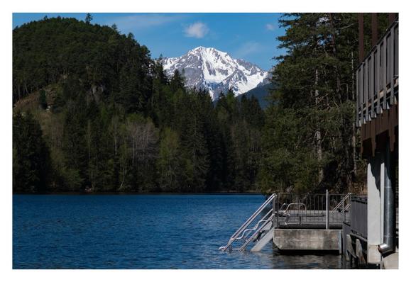 Foto im Querformat. Blick auf einen Bergsee. Rechts ist ein Gebäude angeschnitten im Bild, dahinter eine Terrasse mit einer Leiter, die ins Wasser führt. Der See ist umgeben von dunkelgrünen Nadelbäumen. Am Horizont ragt eine graue, mit Schnee bedeckte Bergspitze über die Bäume, darüber der hellblaue Himmel, an dem einzelne Wölkchen sind. Das Wasser ist dunkelblau, zur Terrasse hin, durch die Sonne, heller blau.