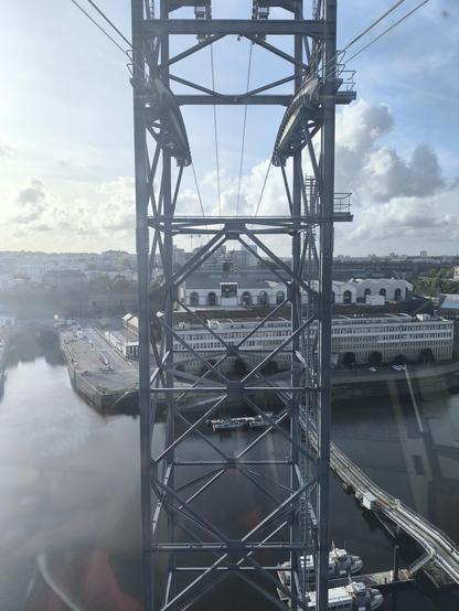 Blick aus der Seilbahn-Kabine auf die Fachwerkstütze in der Mitte des Flusses. Die Stütze ist aus Metall, im oberen Teil sind die Seilspuren zu sehen. In etwas Entfernung sieht man die zweite Kabine.
Unten im Bild ist der Fluss zu sehen.